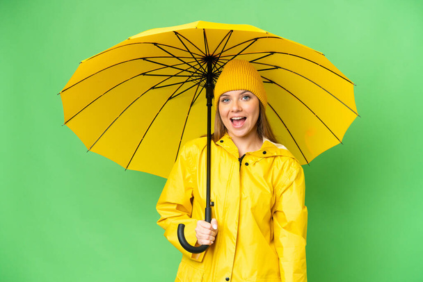 Mulher loira jovem com capa impermeável e guarda-chuva sobre fundo chave croma isolado com expressão facial surpresa - Foto, Imagem