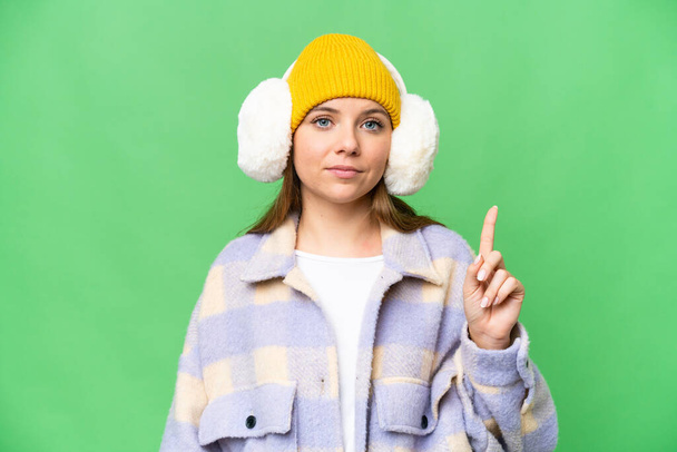 Mujer rubia joven con muffs de invierno sobre fondo de croma clave aislado apuntando con el dedo índice una gran idea - Foto, imagen