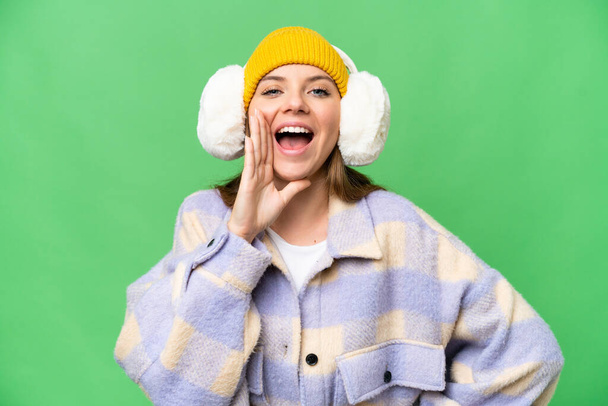 Young blonde woman wearing winter muffs over isolated chroma key background shouting with mouth wide open - Fotó, kép