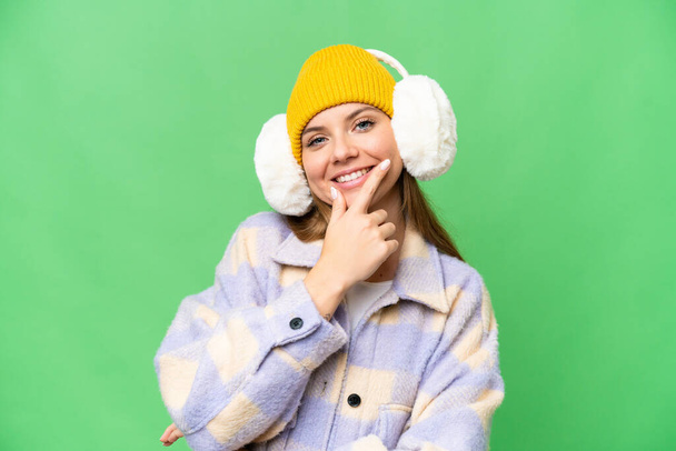Mujer rubia joven con muffs de invierno sobre fondo de croma clave aislado sonriendo - Foto, imagen