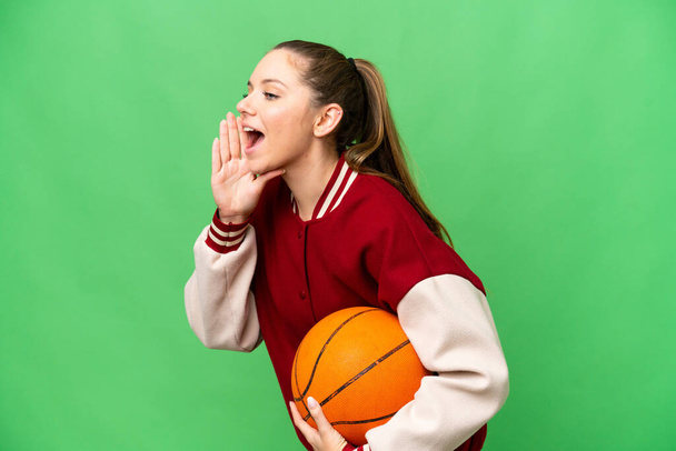 Joven rubia jugando baloncesto sobre fondo de croma key aislado gritando con la boca abierta hacia un lado - Foto, imagen
