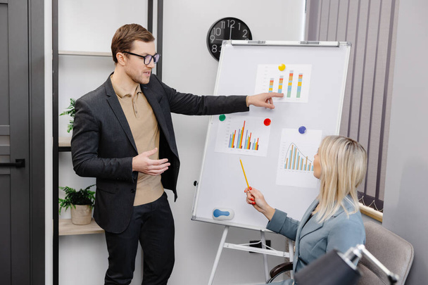 Confident business coach gives presentation on flip chart. A young woman and a man are talking and discussing business project schedules in the office. - Фото, зображення