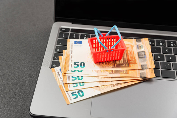 Speelgoed miniatuur winkelwagentje en cash euro op laptop toetsenbord. Concept van online winkelen. Kosten van de boodschappenmand in Europa. - Foto, afbeelding
