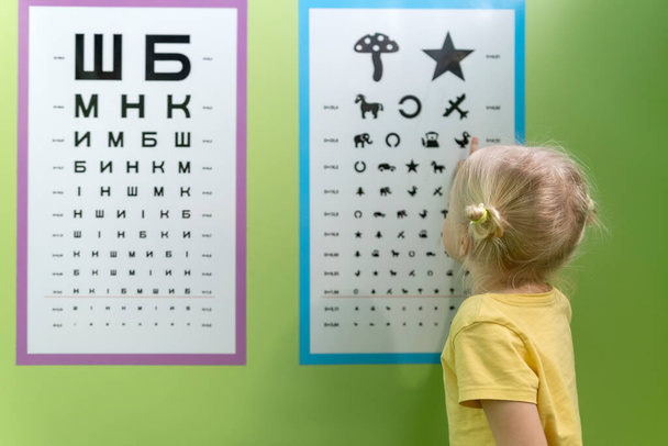 Kleines Mädchen bei einem Termin beim Augenarzt in der Nähe des Posters, um ihr Augenlicht zu überprüfen. Kinderoptometriker für Vorkühlgeräte. - Foto, Bild