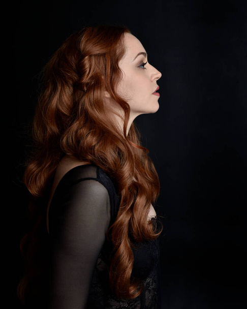Uzun kızıl saçlı, katıksız korse giyen güzel bir kadının portresi. Karanlık stüdyo arka planında karamsar bir şekilde ışıklandırılmış çeşitli yüz ifadeleri.. - Fotoğraf, Görsel
