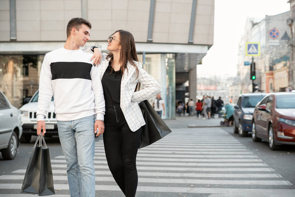 Feliz joven pareja cruza la calle. Chico y chica después de comprar con compras y tiene bolsas de compras en las manos - Foto, imagen