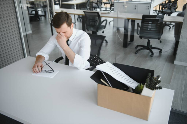 Sad Fired. Let Go Office Worker Packs His Belongings into Cardboard Box and Leaves Office. Workforce Reduction, Downsizing, Reorganization, Restructuring, Outsourcing. Mass Unemployment Market Crisis. - Фото, изображение