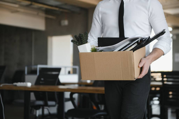 Sad Fired. Let Go Office Worker Packs His Belongings into Cardboard Box and Leaves Office. Workforce Reduction, Downsizing, Reorganization, Restructuring, Outsourcing. Mass Unemployment Market Crisis. - Фото, изображение