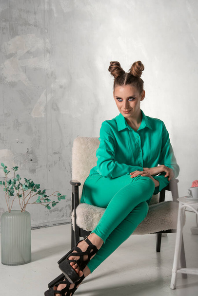 Stylish girl in turquoise shirt and trousers sits elegantly on chair in studio. Model with bun hairstyle and evening makeup poses on gray wall background. - Photo, image