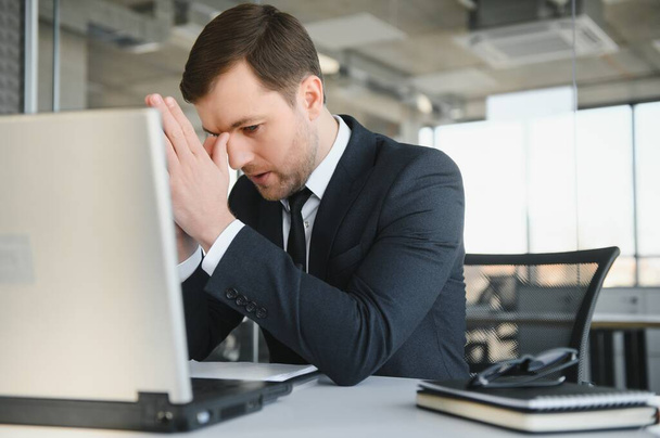 Young stressed handsome businessman working at desk in modern office shouting at laptop screen and being angry about financial situation, jealous of rival capabilities, unable to meet client needs. - Photo, image