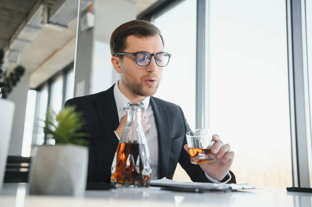 Fatigue young businessman sitting at workplace and holding whiskey bottle, drinking alcohol, bankruptcy concept - Zdjęcie, obraz