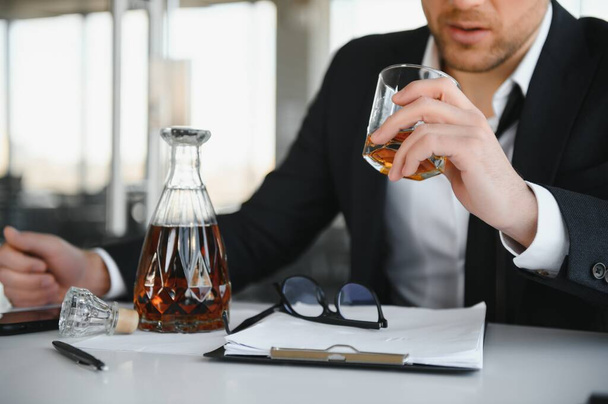 Fatigue young businessman sitting at workplace and holding whiskey bottle, drinking alcohol, bankruptcy concept - 写真・画像