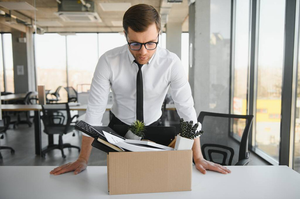 Sad Fired. Let Go Office Worker Packs His Belongings into Cardboard Box and Leaves Office. Workforce Reduction, Downsizing, Reorganization, Restructuring, Outsourcing. Mass Unemployment Market Crisis. - Zdjęcie, obraz