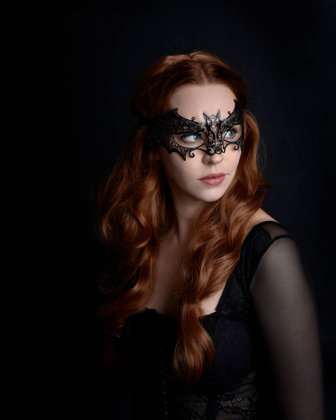 Masked close up portrait of beautiful woman with long red hair wearing sheer corset top, and black bat wing mask.  Isolated on dark studio background with. Moody silhouette lighting. - Valokuva, kuva