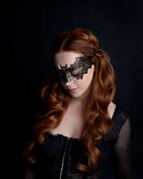 Masked close up portrait of beautiful woman with long red hair wearing sheer corset top, and black bat wing mask.  Isolated on dark studio background with. Moody silhouette lighting. - Zdjęcie, obraz