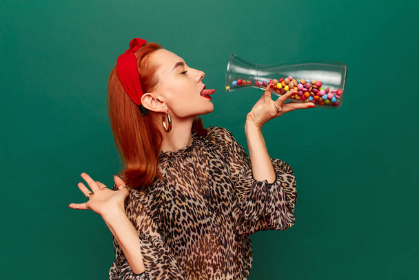 Beautiful young redhead woman in stylish clothes eating multicolored candies over dark green studio background. Concept of food pop art photography. Complementary colors. Copy space for ad, text - Foto, immagini