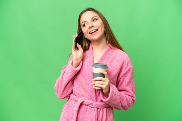 Young woman in a bathrobe over isolated chroma key background holding coffee to take away and a mobile - 写真・画像