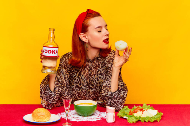 Food pop art photography. Beautiful redhead woman sitting at table against yellow background and having dinner, drinking vodka with onion. Complementary colors. Copy space for ad, text - Фото, зображення
