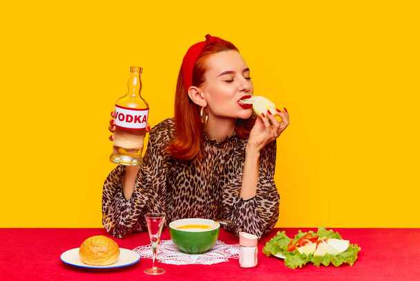 Food pop art photography. Beautiful redhead woman sitting at table against yellow background and having dinner, drinking vodka with onion and salad. Complementary colors. Copy space for ad, text - Photo, Image