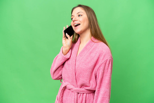 Young woman in a bathrobe over isolated chroma key background keeping a conversation with the mobile phone - Фото, изображение