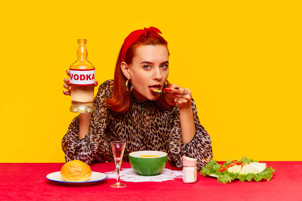 Beautiful stylish redhead woman sitting at table against yellow background and having dinner with cheese soup and drinking vodka. Food pop art photography. Complementary colors. Copy space for ad - Photo, Image