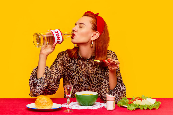 Beautiful redhead woman sitting at table against yellow background and having dinner with cheese, pumpkin soup and drinking vodka. Food pop art photography. Complementary colors. Copy space for ad - Fotó, kép