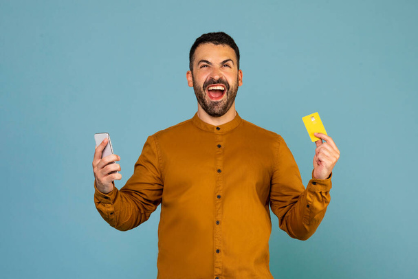 Young emotional man using cellphone and holding credit bank card doing online shopping and celebrating big sale isolated on blue background studio shot - Фото, изображение