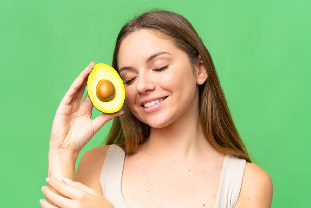 Mulher loira jovem sobre fundo chave croma isolado segurando um abacate enquanto sorri. Fechar retrato - Foto, Imagem