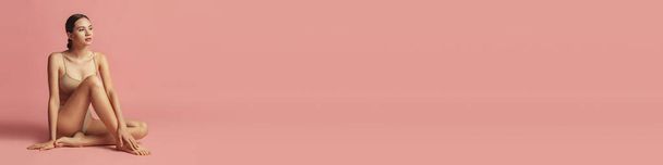 Uma jovem menina bronzeada bonita com figura perfeita em roupa interior deitada no chão sobre fundo de estúdio rosa. Conceito de beleza feminina, ternura, cuidados com o corpo e a pele. Banner, panfleto - Foto, Imagem
