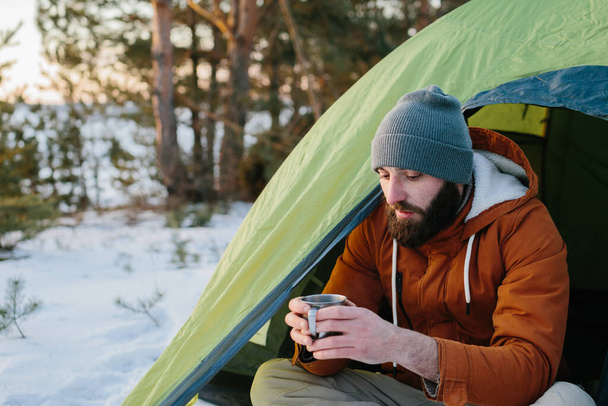 若い髭を生やした男がテントの近くの冬の山に腰を下ろしている。帽子に髭を生やした旅行者と暖かいジャケットは、ハイキングの後に熱いお茶やコーヒーを飲むことによって暖まります。旅行、ライフスタイル - 写真・画像