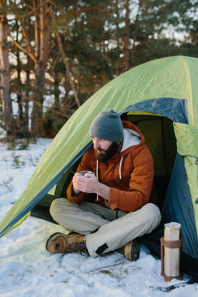 Un joven barbudo descansa en las montañas de invierno cerca de una tienda de campaña. Un hombre viajero con una barba en una gorra y una chaqueta caliente se calienta bebiendo té caliente o café después de una caminata. Viajes, estilo de vida - Foto, imagen