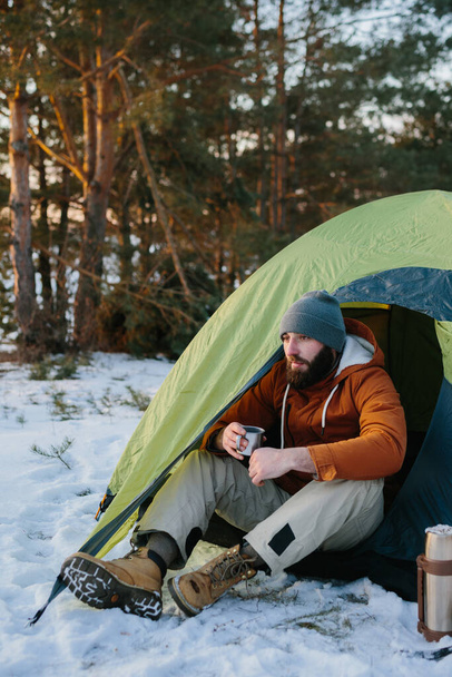 Nuori parrakas mies lepää talvivuorilla lähellä telttaa. Mies-matkustaja, jolla on parta lakissa ja lämmin takki, lämpenee juomalla kuumaa teetä tai kahvia vaelluksen jälkeen. Matkustaminen, elämäntapa - Valokuva, kuva