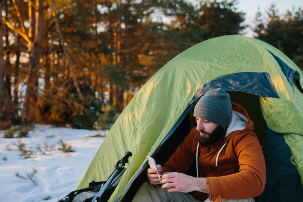 Un turista cansado descansa después de una difícil escalada sentado en una tienda de campaña en un bosque nevado. Un hombre usa su teléfono para enviar un mensaje o revisar su GPS. - Foto, imagen