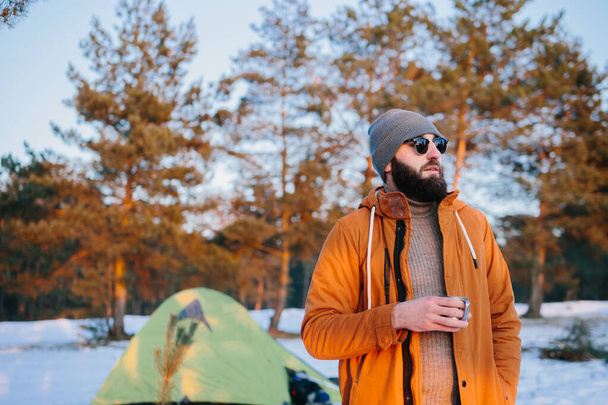 Turisti seisoo lähellä telttaa kupin teetä tai kahvia lämpimässä auringonlaskussa talvimetsässä.. - Valokuva, kuva