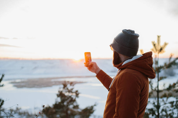 A túrázó élvezi a naplementét a téli hegyekben, és készít egy fényképet a telefonján.. - Fotó, kép