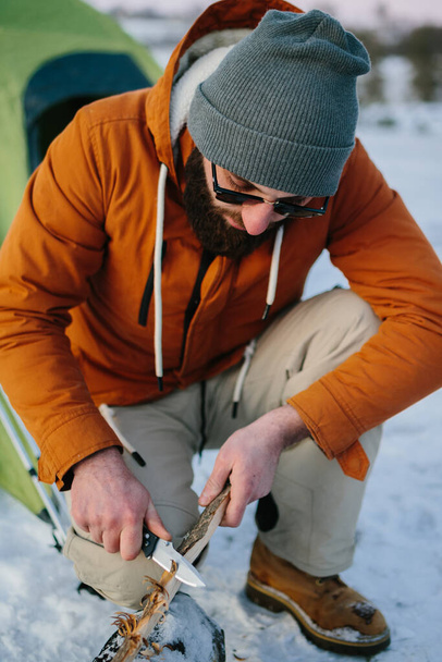 A man prepares wood wool with a knife for starting a fire in a winter forest at sunset. Winter survival concept. - Photo, Image