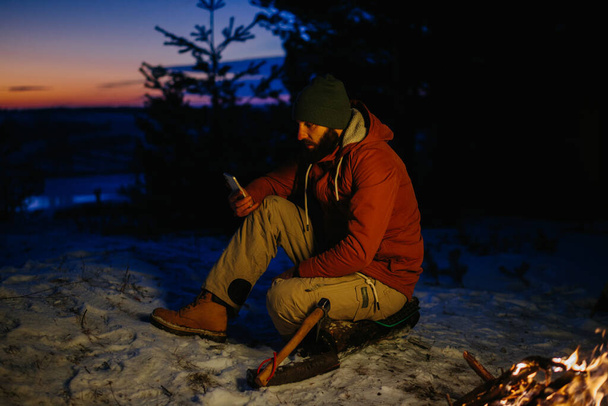 Un excursionista revisa su teléfono mientras se calienta junto a una fogata por la noche en las montañas. - Foto, imagen
