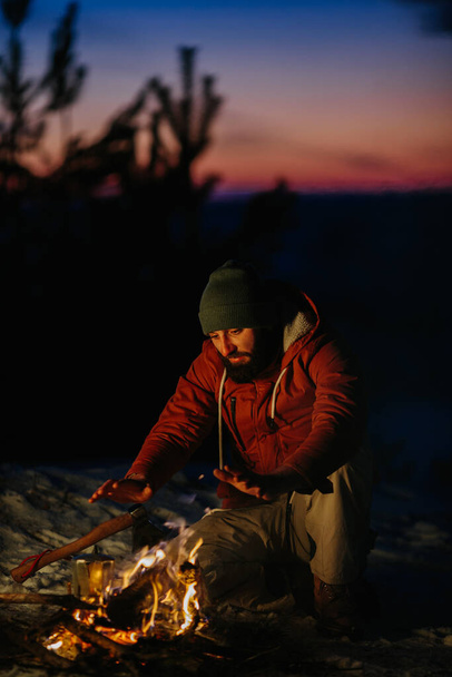 Un hombre se sienta cerca de una fogata, calentándose junto al fuego en un bosque de invierno al atardecer. - Foto, imagen