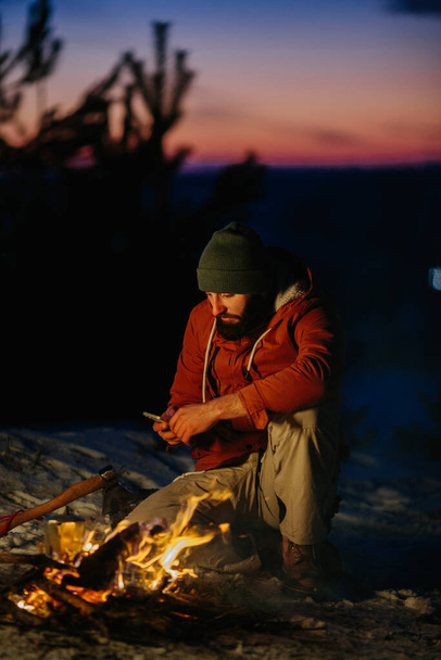 Un excursionista revisa su teléfono mientras se calienta junto a una fogata por la noche en las montañas. - Foto, imagen