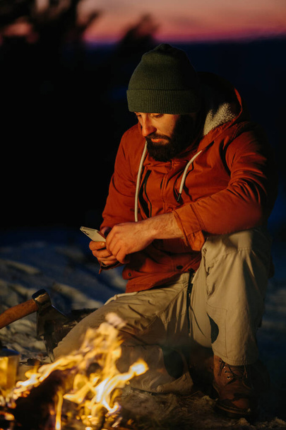 Yürüyüşçünün biri, geceleri dağlarda kamp ateşinde kendini ısıtırken telefonunu kontrol eder.. - Fotoğraf, Görsel