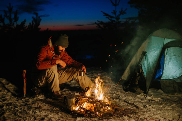 Un joven barbudo descansa junto a la fogata nocturna en el bosque invernal. El concepto de supervivencia y senderismo en la naturaleza. - Foto, imagen