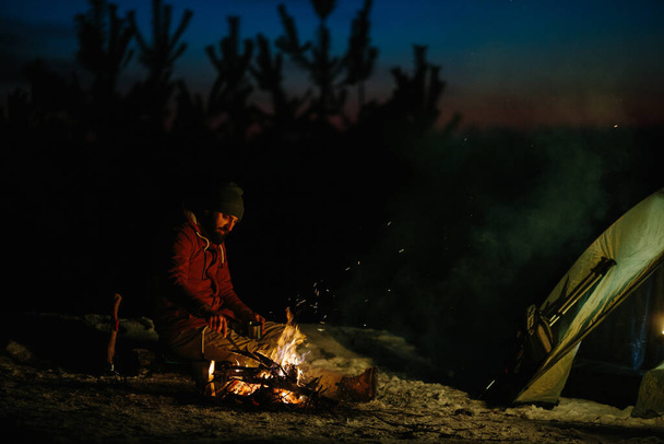 Un joven barbudo hace un delicioso café en una hoguera mientras disfruta de la puesta de sol en un bosque de invierno. El concepto de supervivencia y senderismo en la naturaleza. - Foto, imagen