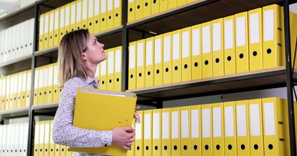Przemyślana, skoncentrowana kobieta patrzy na szafkę z żółtymi teczkami dokumentów. Archiwum i dokumentacja biznesowa - Materiał filmowy, wideo