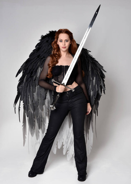 Full length portrait of beautiful woman with long red hair wearing  corset top, leather pants, large black angel feather wings. Standing pose holding sword weapon, walking forwards with gestural hands reaching out. Isolated on white studio background - Fotó, kép