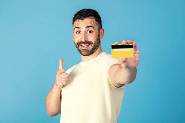 Glücklicher Mann zeigt auf gelbe Kreditkarte in der Hand und lächelt, genießt unbegrenzten Einkauf und Online-Bezahlung, blauer Studiohintergrund - Foto, Bild