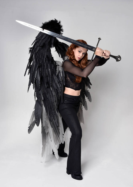 Full length portrait of beautiful woman with long red hair wearing  corset top, leather pants, large black angel feather wings. Standing pose holding sword weapon, walking forwards with gestural hands reaching out. Isolated on white studio background - 写真・画像