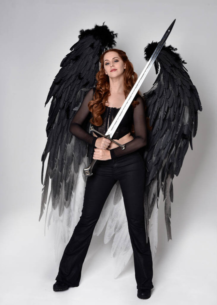 Full length portrait of beautiful woman with long red hair wearing  corset top, leather pants, large black angel feather wings. Standing pose holding sword weapon, walking forwards with gestural hands reaching out. Isolated on white studio background - Fotoğraf, Görsel