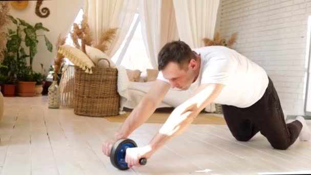 Athletic man is doing exercises with an abs wheel in living room at home. Sport and fitness. Workout and wellness concept. Fitness workout of guy. High-intensity interval training. - 映像、動画