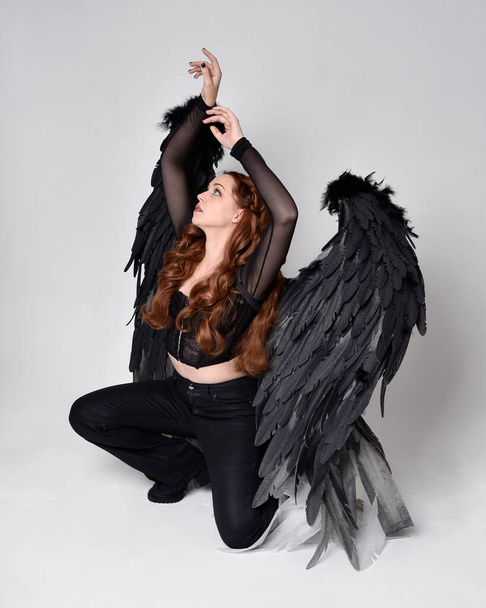 Full length portrait of beautiful woman with long red hair wearing sheer corset top, leather pants & large black angel feather wings.  Sitting pose with gestural hands reaching out, kneeling on floor. Isolated on white studio background - Fotó, kép