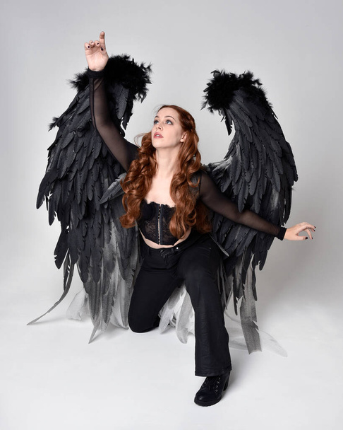 Full length portrait of beautiful woman with long red hair wearing sheer corset top, leather pants & large black angel feather wings.  Sitting pose with gestural hands reaching out, kneeling on floor. Isolated on white studio background - Foto, immagini